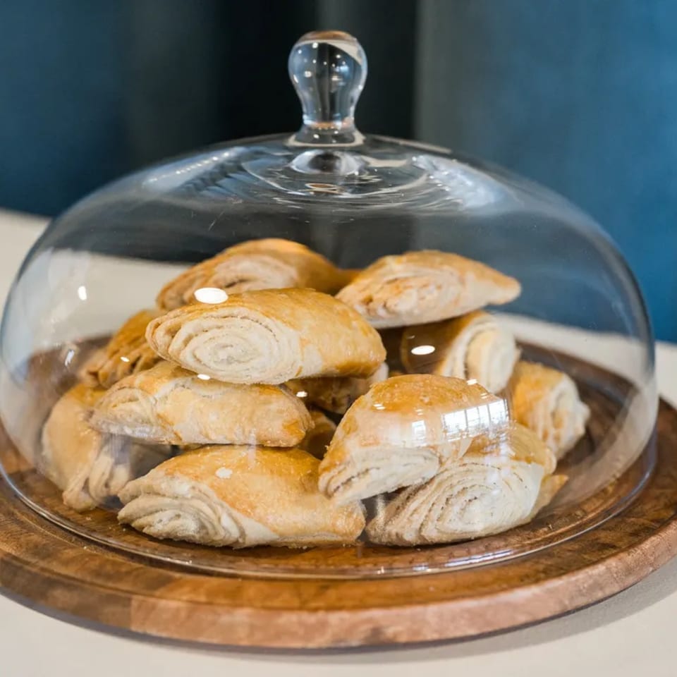 Golden-brown pastries, possibly Mediterranean, are displayed under a clear glass cloche on a wooden tray.