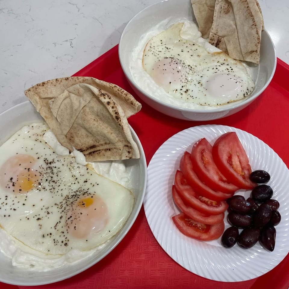 Two bowls of fried eggs with yogurt and pita, alongside sliced tomatoes and olives on a red tray.
