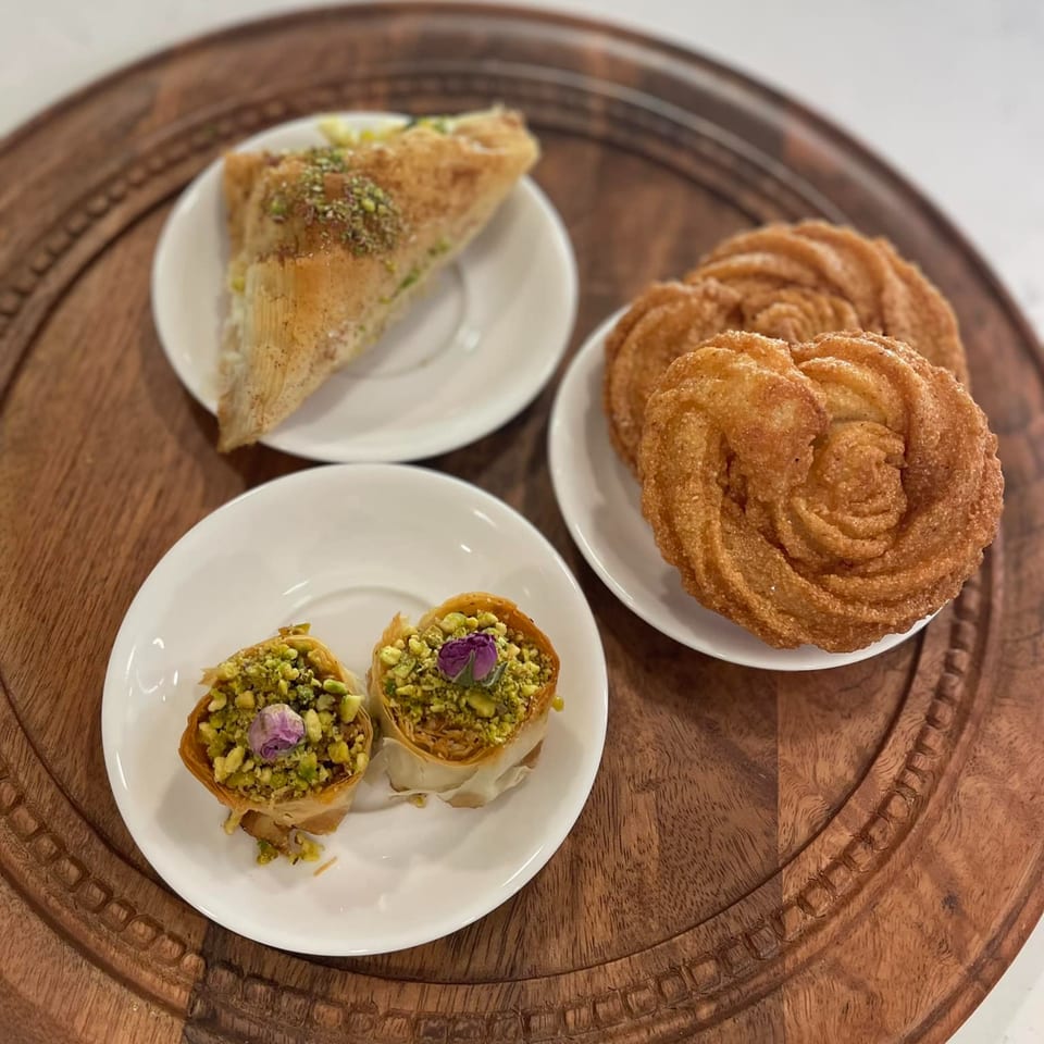 A wooden tray holds three plates of Mediterranean desserts: baklava, pistachio bird's nests, and churro-like pastries.
