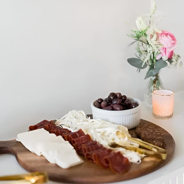 A Mediterranean charcuterie board with cheeses, cured meats, olives, and bread, next to a floral arrangement.