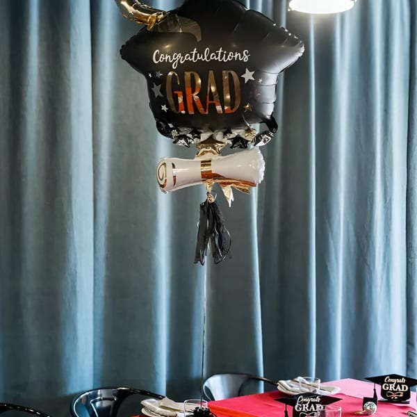 A black graduation balloon centerpiece with a diploma balloon and tassels on a red tablecloth.