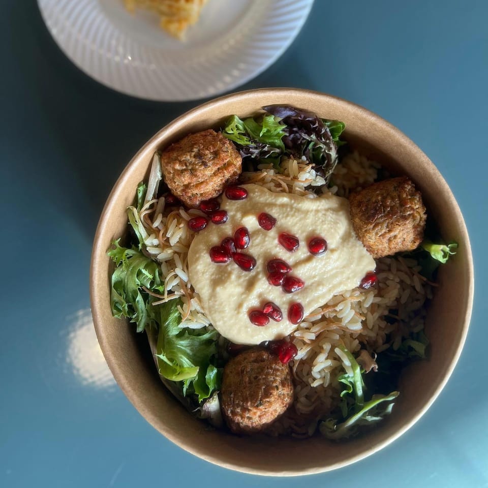 Mediterranean bowl with rice, falafel, hummus, pomegranate seeds, and mixed greens in a paper bowl.