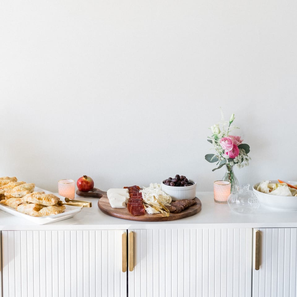 A Mediterranean spread with pastries, charcuterie, olives, and a floral arrangement on a white counter.