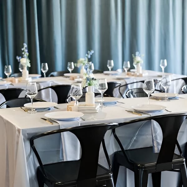 Tables set with white tablecloths, plates, glasses, and floral centerpieces against blue curtains.