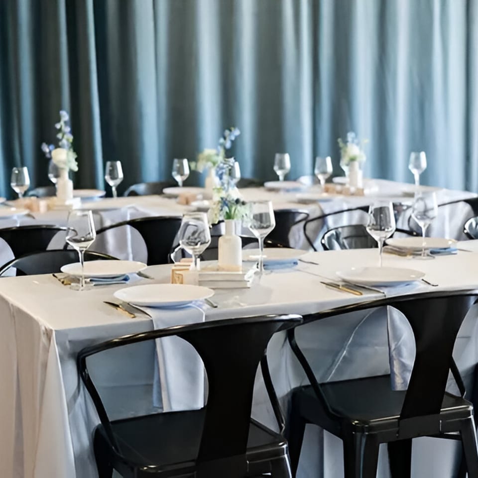 Tables set with white tablecloths, plates, glasses, and floral centerpieces against blue curtains.