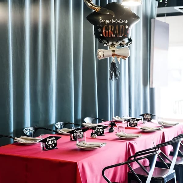 A Cafe POM table set for a graduation party with a pink tablecloth and "Congrats Grad" decorations.