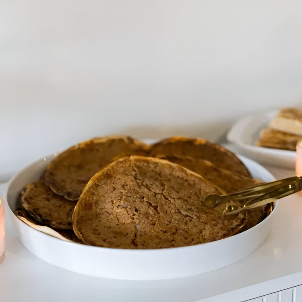 A white bowl filled with several round, brown flatbreads, with tongs resting on the side.