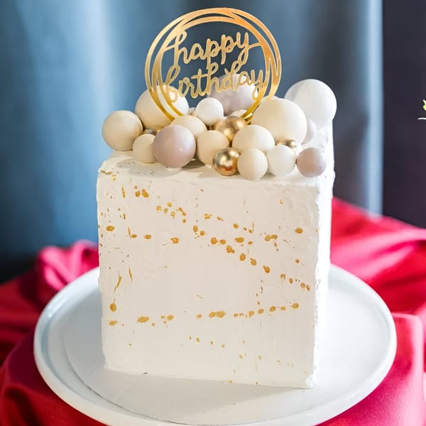White square birthday cake with gold speckles, topped with white and gold spheres and a "Happy Birthday" topper.